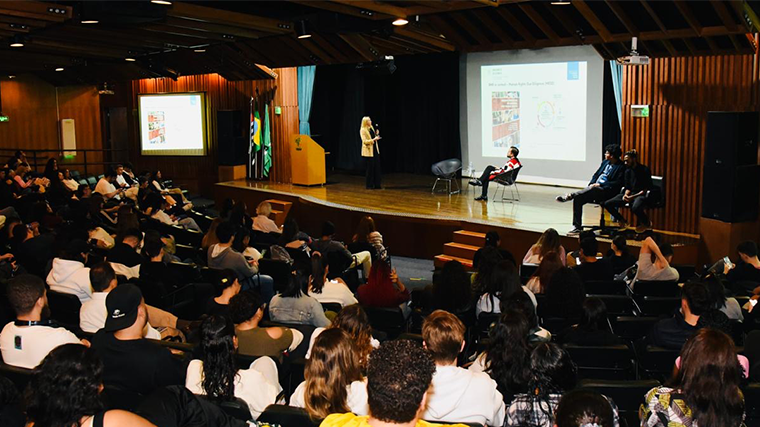 A speaker addresses an audience in a large lecture hall, with three panelists seated on stage and a presentation displayed on a screen